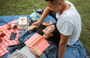 man and woman picnicing woman laying back with book on her chest smiling at male companion